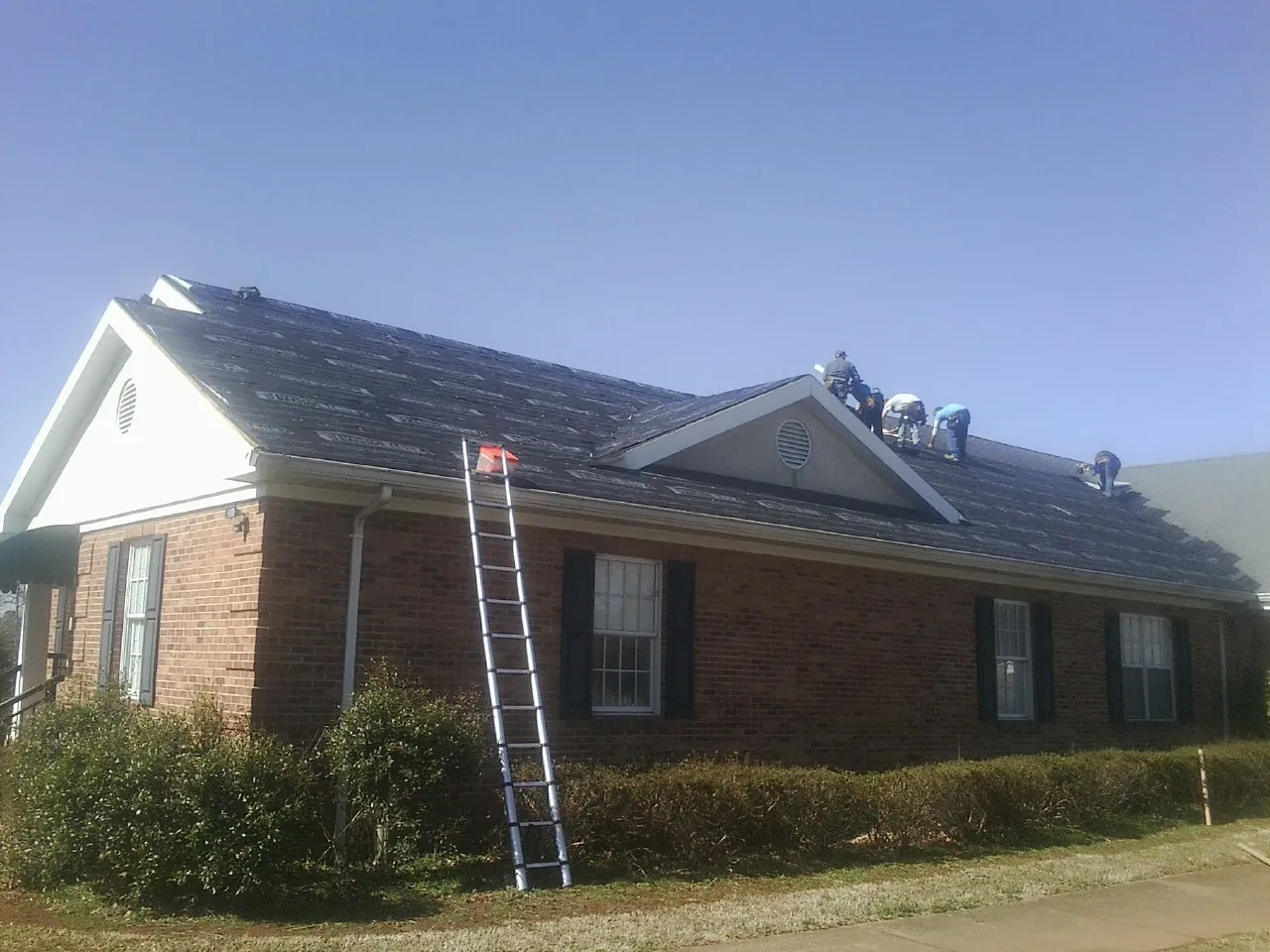 Crew tearing shingles off a brick ranch roof with a ladder set — Emergency Roof Repair work by our crew in Queensbury