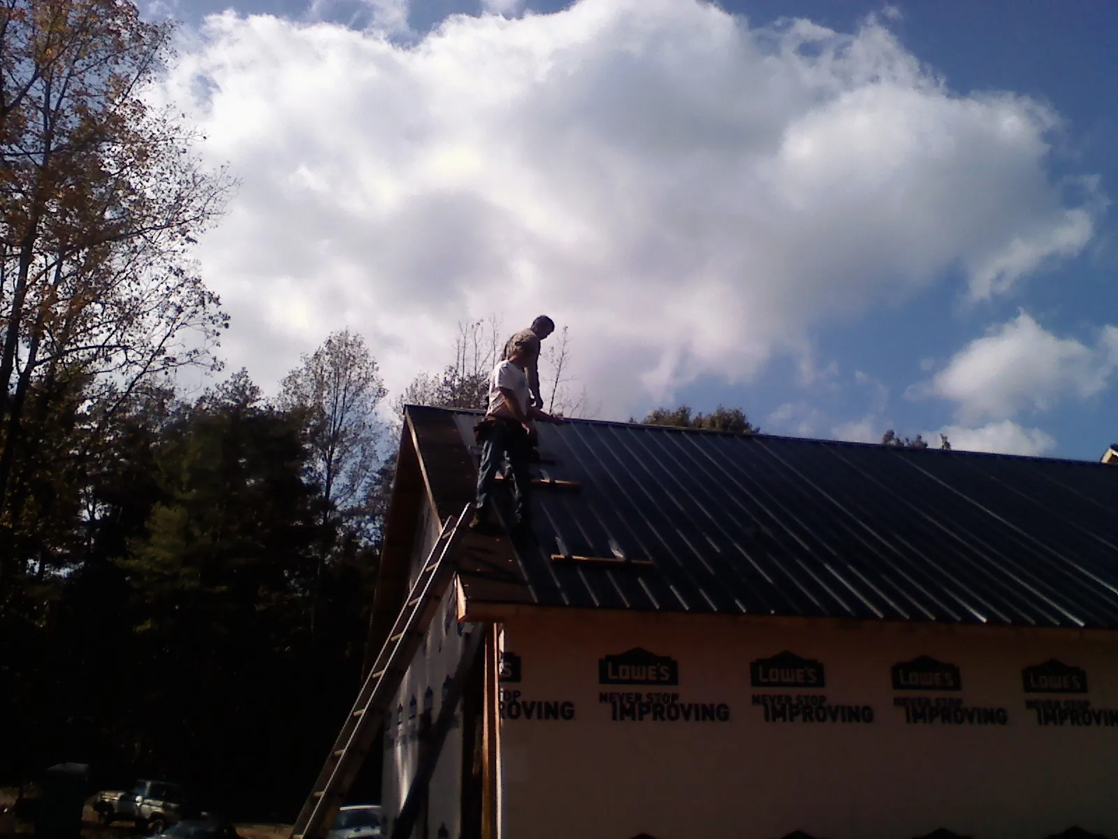 Roofer fastening dark metal panels high against a cloudy sky, from a Roof Inspection job in Queensbury